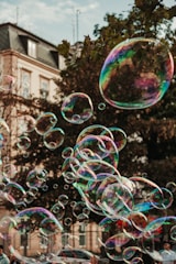 Numerous soap bubbles of varying sizes float through the air against the backdrop of a building and trees. The bubbles are iridescent, reflecting multiple colors such as pink, blue, and green. In the foreground, partially visible hands appear to be reaching towards the bubbles.