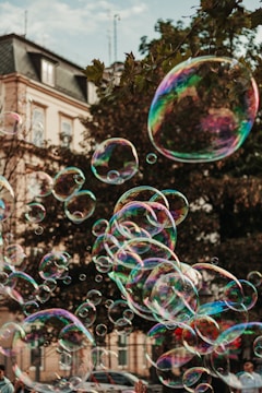 Numerous soap bubbles of varying sizes float through the air against the backdrop of a building and trees. The bubbles are iridescent, reflecting multiple colors such as pink, blue, and green. In the foreground, partially visible hands appear to be reaching towards the bubbles.