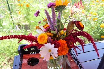 A vibrant bouquet of diverse flowers arranged in a glass vase. The bouquet includes sunflowers, marigolds, daisies, and other colorful blooms with varying textures and shapes. The flowers rest on a wooden table with a garden full of blossoming flowers in the blurred background.