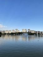 A serene lakeside scene with a line of tall residential buildings in the background reflecting on the calm water. Multiple fountains create arcs of water in the lake. A clear blue sky stretches above, contrasting with the greenery and urban landscape.