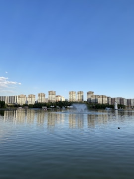 A serene lakeside scene with a line of tall residential buildings in the background reflecting on the calm water. Multiple fountains create arcs of water in the lake. A clear blue sky stretches above, contrasting with the greenery and urban landscape.