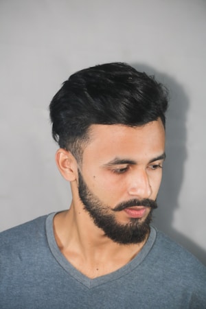 A man with a neatly groomed beard and mustache is looking down to his right. He has short black hair and is wearing a gray V-neck t-shirt. The background is plain and light-colored, creating a subtle contrast with his dark hair.