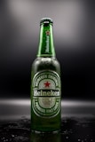 A Heineken beer bottle stands upright against a dark background, with droplets of condensation visible on the green glass. The label features the brand's logo and a red star, with text indicating it is a premium malt lager brewed in Holland.