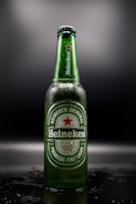 A Heineken beer bottle stands upright against a dark background, with droplets of condensation visible on the green glass. The label features the brand's logo and a red star, with text indicating it is a premium malt lager brewed in Holland.