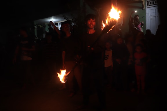 A person is performing a fire dance surrounded by a crowd. The performer holds flaming objects and is the focal point of the scene. The background shows a mix of adults and children watching attentively, some capturing the moment with their phones. The setting appears to be outdoors at night in a dimly lit area.