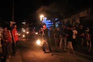 A group of people gather on a dimly lit street at night. One individual stands in the center, performing with fire, holding flaming objects in their hands. The crowd, including both men and women, watches the performance, some with phones in hand. The surrounding buildings and ambient darkness create an atmospheric setting.