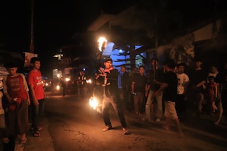 A group of people gather on a dimly lit street at night. One individual stands in the center, performing with fire, holding flaming objects in their hands. The crowd, including both men and women, watches the performance, some with phones in hand. The surrounding buildings and ambient darkness create an atmospheric setting.