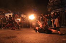 A nighttime street performance with a fire-breathing act is taking place. Two performers are engaged in dynamic movements on the ground while holding burning torches. A crowd gathers around, watching attentively, with expressions of intrigue and amazement.