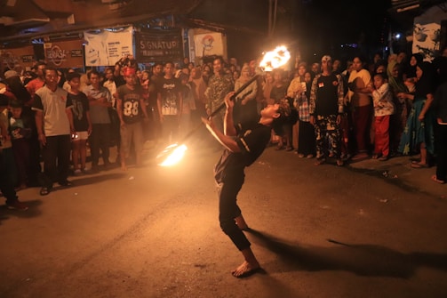 A performer skillfully breathes fire amidst a street crowd watching attentively. The scene is illuminated by the flames, casting a warm glow on the audience, who are standing in a circle.