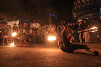 A street performance featuring two performers engaging in a fire dance routine. One performer is balancing on their hands with flames visible on both ends of a stick, while a crowd of people watches them in the background. The scene is set outdoors during nighttime, contributing to the dramatic effect of the fire.