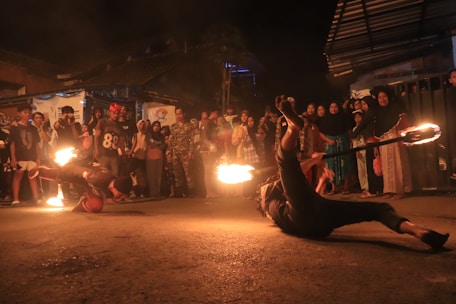 A street performance featuring two performers engaging in a fire dance routine. One performer is balancing on their hands with flames visible on both ends of a stick, while a crowd of people watches them in the background. The scene is set outdoors during nighttime, contributing to the dramatic effect of the fire.