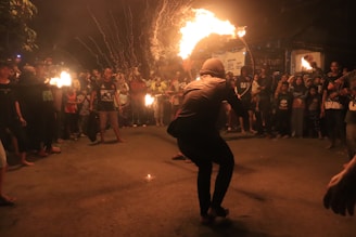 A street performance taking place at night, featuring a person skillfully handling a flaming object while surrounded by a crowd. The audience is gathered around attentively watching the performer in the center. The scene is lively with bright flames illuminating the surroundings against the dark backdrop.