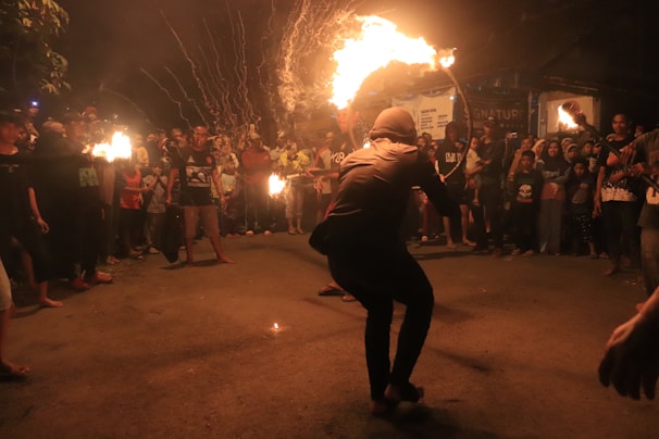 A street performance taking place at night, featuring a person skillfully handling a flaming object while surrounded by a crowd. The audience is gathered around attentively watching the performer in the center. The scene is lively with bright flames illuminating the surroundings against the dark backdrop.