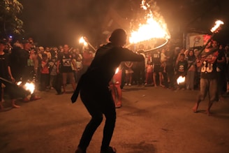 A group of people gathered outdoors at night, watching a fire performance. The performer is holding a flaming object and surrounded by an audience standing in a circle. The background is dimly lit with a mix of shadows and glowing firelight.