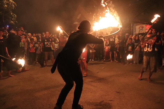 A group of people gathered outdoors at night, watching a fire performance. The performer is holding a flaming object and surrounded by an audience standing in a circle. The background is dimly lit with a mix of shadows and glowing firelight.