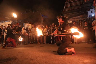 A night-time street performance features two performers engaging in a fire dance, surrounded by an attentive crowd. The performers are kneeling, holding blazing torches, and creating trails of fire as they move. The audience consists of people of various ages, standing closely together, watching the performance with interest. The setting is outdoors, with dim lighting highlighting the orange glow of the fire.
