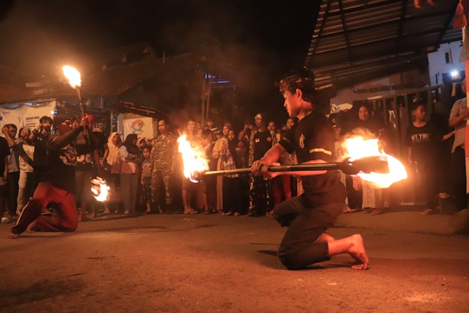A night-time street performance features two performers engaging in a fire dance, surrounded by an attentive crowd. The performers are kneeling, holding blazing torches, and creating trails of fire as they move. The audience consists of people of various ages, standing closely together, watching the performance with interest. The setting is outdoors, with dim lighting highlighting the orange glow of the fire.