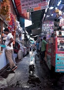 A bustling market alley filled with various stalls selling mobile accessories. Colorful garlands hang from the roof, and signboards for brands like Samsung, Oppo, and iPhone are visible. Shoppers and vendors are engaged in transactions.