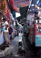A bustling market alley filled with various stalls selling mobile accessories. Colorful garlands hang from the roof, and signboards for brands like Samsung, Oppo, and iPhone are visible. Shoppers and vendors are engaged in transactions.