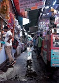 A bustling market alley filled with various stalls selling mobile accessories. Colorful garlands hang from the roof, and signboards for brands like Samsung, Oppo, and iPhone are visible. Shoppers and vendors are engaged in transactions.
