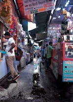 A bustling market alley filled with various stalls selling mobile accessories. Colorful garlands hang from the roof, and signboards for brands like Samsung, Oppo, and iPhone are visible. Shoppers and vendors are engaged in transactions.