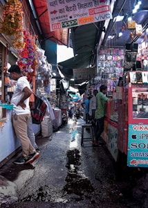 A bustling market alley filled with various stalls selling mobile accessories. Colorful garlands hang from the roof, and signboards for brands like Samsung, Oppo, and iPhone are visible. Shoppers and vendors are engaged in transactions.