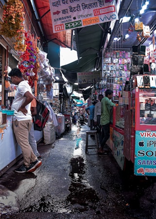 A bustling market alley filled with various stalls selling mobile accessories. Colorful garlands hang from the roof, and signboards for brands like Samsung, Oppo, and iPhone are visible. Shoppers and vendors are engaged in transactions.