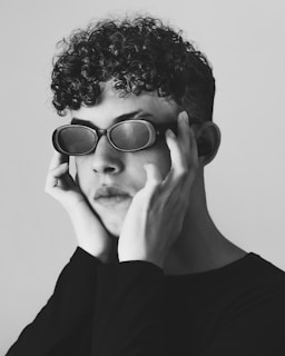 A black-and-white portrait of a person with curly hair wearing oval-shaped sunglasses. The individual's hands are positioned around their face, lightly touching the edges of the sunglasses. The expression appears contemplative, and the composition emphasizes the texture of the hair and the smoothness of the skin.