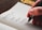 A hand holding a pen writes a list in a notebook. The page is lined and contains entries such as Household Bills, Power, Internet, Insurance, and Groceries. The background features a wooden table and a few colorful objects.