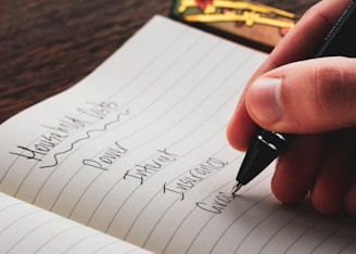 A hand holding a pen writes a list in a notebook. The page is lined and contains entries such as Household Bills, Power, Internet, Insurance, and Groceries. The background features a wooden table and a few colorful objects.