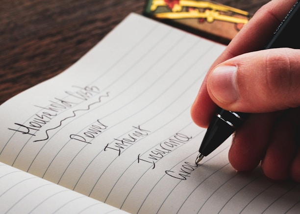 A hand holding a pen writes a list in a notebook. The page is lined and contains entries such as Household Bills, Power, Internet, Insurance, and Groceries. The background features a wooden table and a few colorful objects.