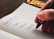 A hand holding a pen writes a list in a notebook. The page is lined and contains entries such as Household Bills, Power, Internet, Insurance, and Groceries. The background features a wooden table and a few colorful objects.