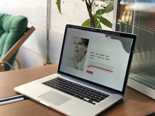 A laptop is open on a wooden table, displaying a webpage with text and a black-and-white photo of a smiling woman. In the background, there is a green chair and a leafy plant against a wall.