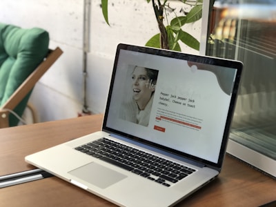 A laptop is open on a wooden table, displaying a webpage with text and a black-and-white photo of a smiling woman. In the background, there is a green chair and a leafy plant against a wall.