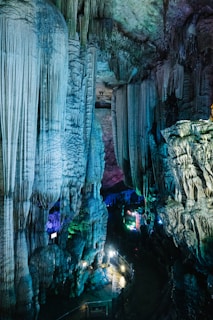 A large cave interior featuring prominent stalactites and stalagmites illuminated by colorful artificial lights, creating a surreal and mystical atmosphere. The cave walls and formations showcase intricate textures and patterns, with pathways allowing for exploration.