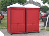 Two red portable toilets are positioned side by side on a paved area outdoors. Nearby, a wooden gazebo and several vehicles are visible, including a red truck. A person is sitting on the grass to the left, with trees and other structures in the background.