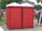 Two red portable toilets are positioned side by side on a paved area outdoors. Nearby, a wooden gazebo and several vehicles are visible, including a red truck. A person is sitting on the grass to the left, with trees and other structures in the background.