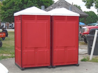 Two red portable toilets are positioned side by side on a paved area outdoors. Nearby, a wooden gazebo and several vehicles are visible, including a red truck. A person is sitting on the grass to the left, with trees and other structures in the background.