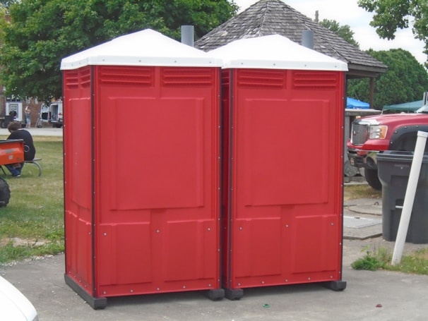 Two red portable toilets are positioned side by side on a paved area outdoors. Nearby, a wooden gazebo and several vehicles are visible, including a red truck. A person is sitting on the grass to the left, with trees and other structures in the background.