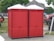 Two red portable toilets are positioned side by side on a paved area outdoors. Nearby, a wooden gazebo and several vehicles are visible, including a red truck. A person is sitting on the grass to the left, with trees and other structures in the background.
