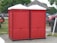 Two red portable toilets are positioned side by side on a paved area outdoors. Nearby, a wooden gazebo and several vehicles are visible, including a red truck. A person is sitting on the grass to the left, with trees and other structures in the background.