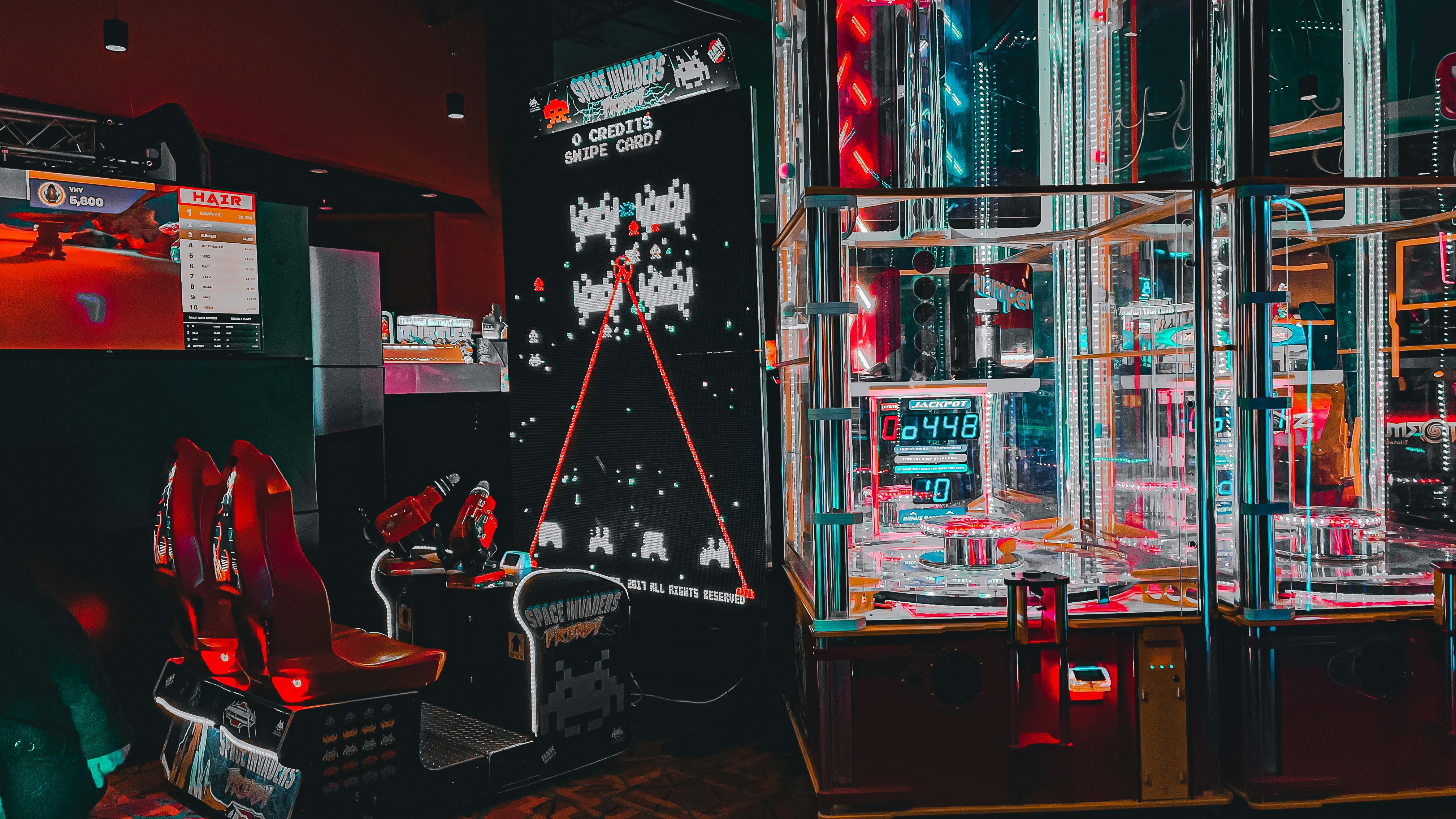 An arcade setting featuring a classic Space Invaders game setup and a modern gaming machine with seats. Bright neon lights illuminate the area, with scores displayed on digital screens. A bright and visually engaging environment full of electronic components.