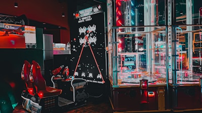 An arcade setting featuring a classic Space Invaders game setup and a modern gaming machine with seats. Bright neon lights illuminate the area, with scores displayed on digital screens. A bright and visually engaging environment full of electronic components.