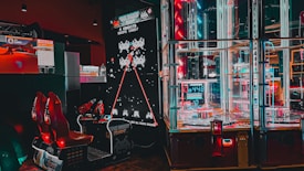 An arcade setting featuring a classic Space Invaders game setup and a modern gaming machine with seats. Bright neon lights illuminate the area, with scores displayed on digital screens. A bright and visually engaging environment full of electronic components.
