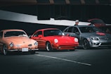 Three parked cars, a peach-colored vintage car, a red sports car, and a gray modern sedan are displayed in a parking area. A person wearing a hat stands beside the gray car, which has its trunk open. The cars are set against a backdrop of a large, dark-colored vehicle, possibly a bus.