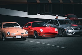 Three parked cars, a peach-colored vintage car, a red sports car, and a gray modern sedan are displayed in a parking area. A person wearing a hat stands beside the gray car, which has its trunk open. The cars are set against a backdrop of a large, dark-colored vehicle, possibly a bus.
