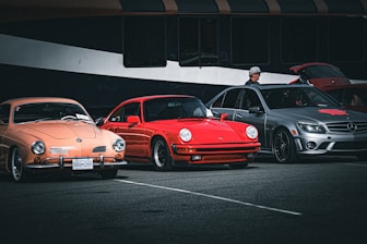 Three parked cars, a peach-colored vintage car, a red sports car, and a gray modern sedan are displayed in a parking area. A person wearing a hat stands beside the gray car, which has its trunk open. The cars are set against a backdrop of a large, dark-colored vehicle, possibly a bus.