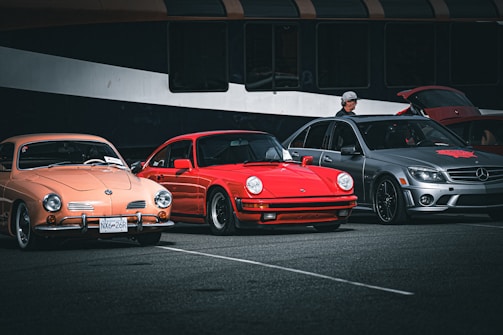 Three parked cars, a peach-colored vintage car, a red sports car, and a gray modern sedan are displayed in a parking area. A person wearing a hat stands beside the gray car, which has its trunk open. The cars are set against a backdrop of a large, dark-colored vehicle, possibly a bus.