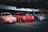 Three parked cars, a peach-colored vintage car, a red sports car, and a gray modern sedan are displayed in a parking area. A person wearing a hat stands beside the gray car, which has its trunk open. The cars are set against a backdrop of a large, dark-colored vehicle, possibly a bus.