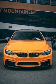 A bright orange sports car is parked in front of a large, blue and gold train. The train bears the name 'Rocky Mountaineer' and has multiple windows visible. The car has a sleek design, prominent grille, and is positioned straight on, occupying the foreground of the image.